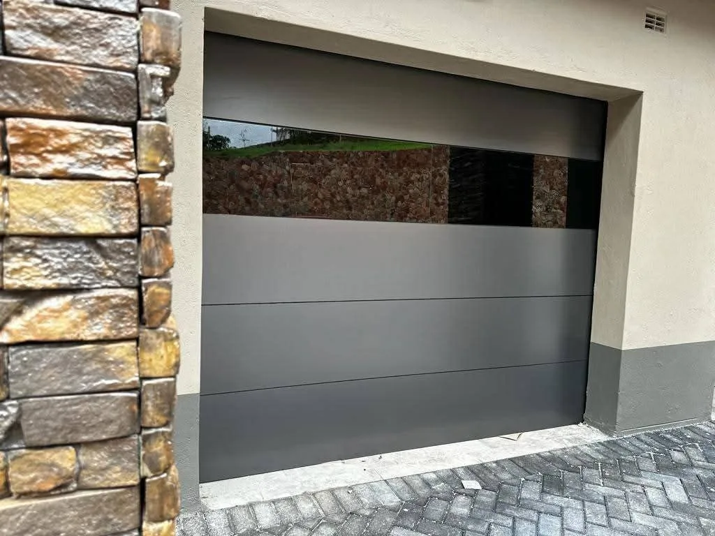 A grey sectional garage door with a wide glazed panel beside a stone-clad wall.