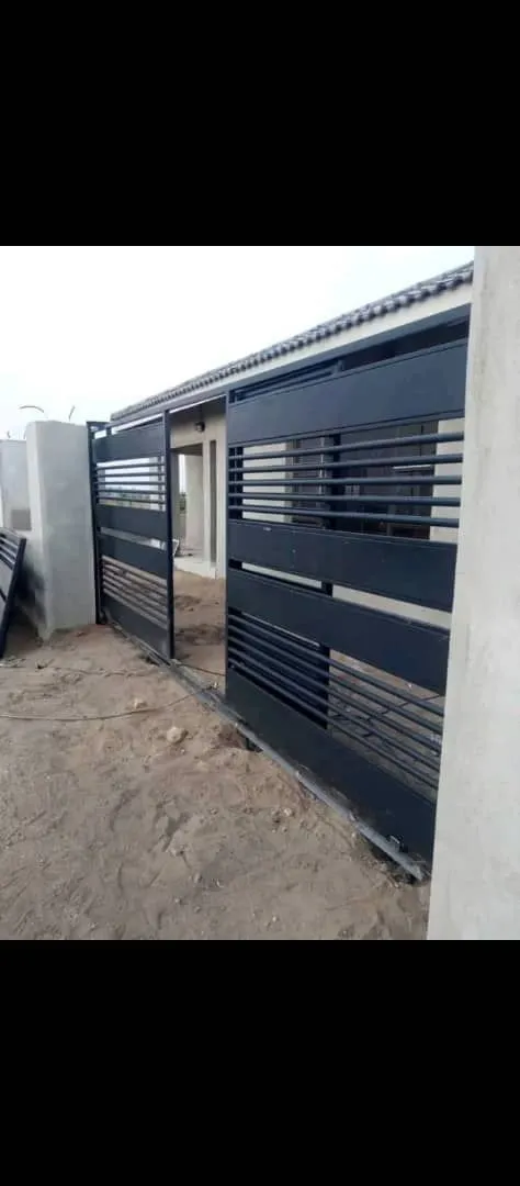 A dark slatted gate with a pedestrian opening during fitting beside a plastered boundary wall.