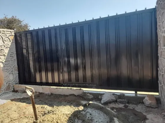 A black vertical-panel driveway gate between stone-clad pillars, with groundworks still visible.